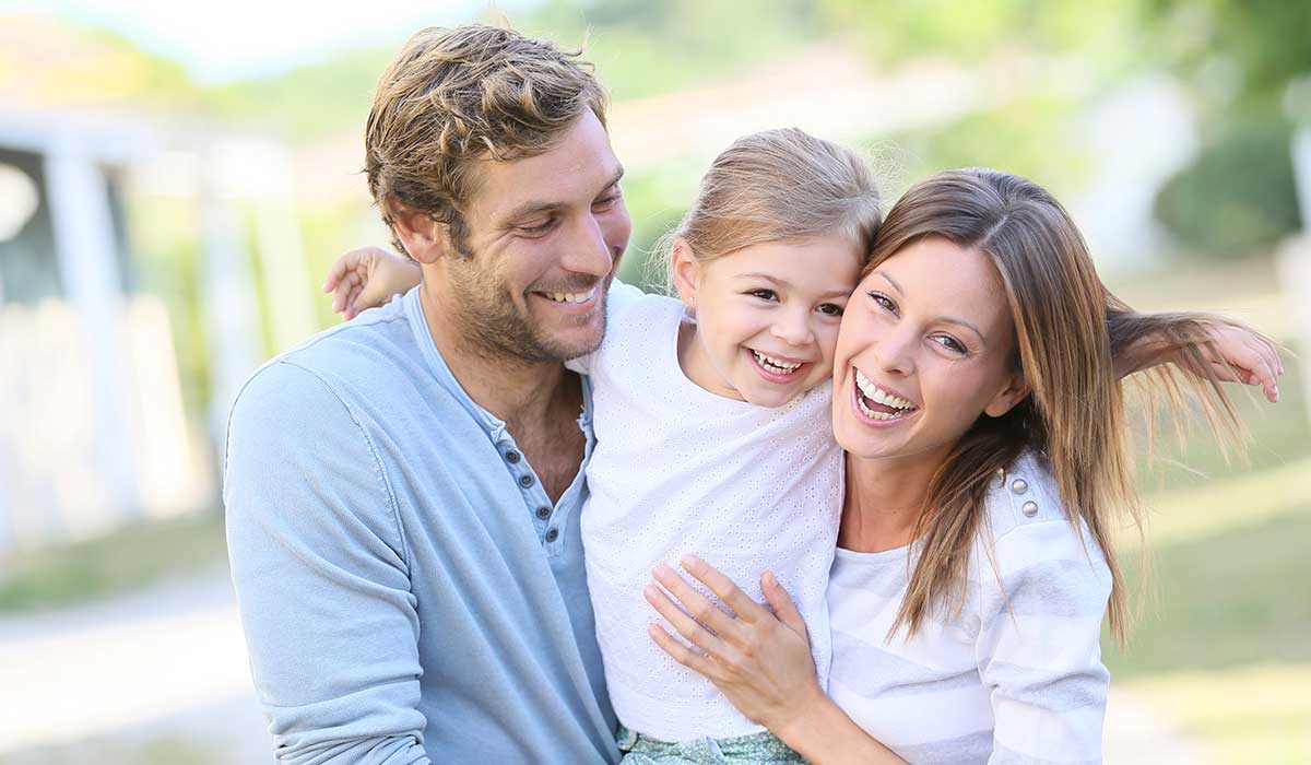 A happy family of three—an adult man, an adult woman, and a young girl—smiles and embraces each other outdoors on a sunny day. The girl is between the adults, who both look joyful and relaxed.