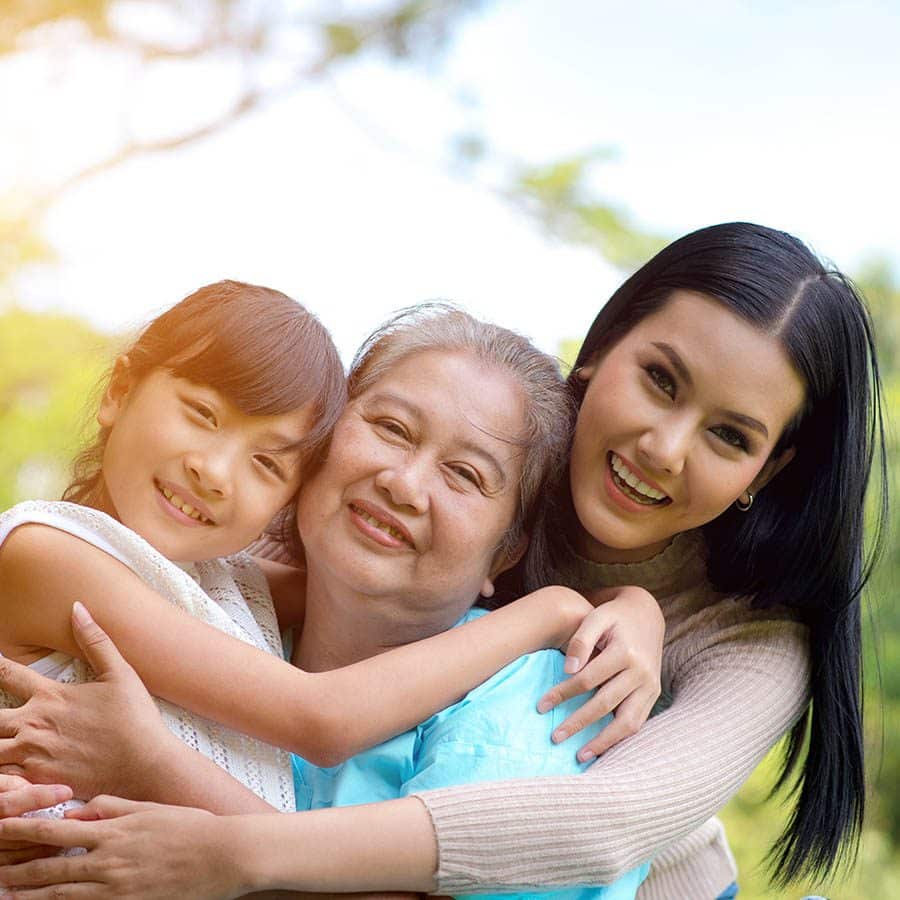 Three generations of women‚Äîchild, elderly woman, and adult‚Äîembrace each other and smile warmly outdoors, with trees and soft sunlight in the background.