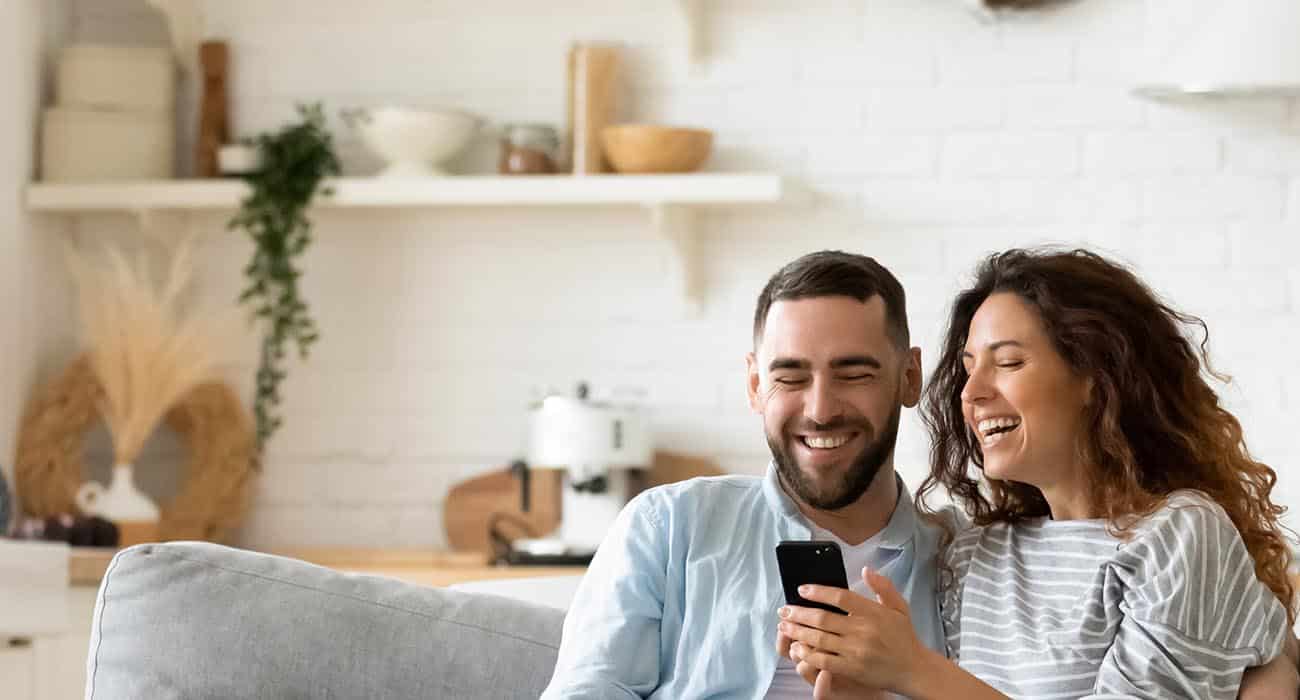 A smiling couple sits on a couch at home, looking at a smartphone together. The background shows a bright, cozy kitchen with shelves, plants, and kitchenware.